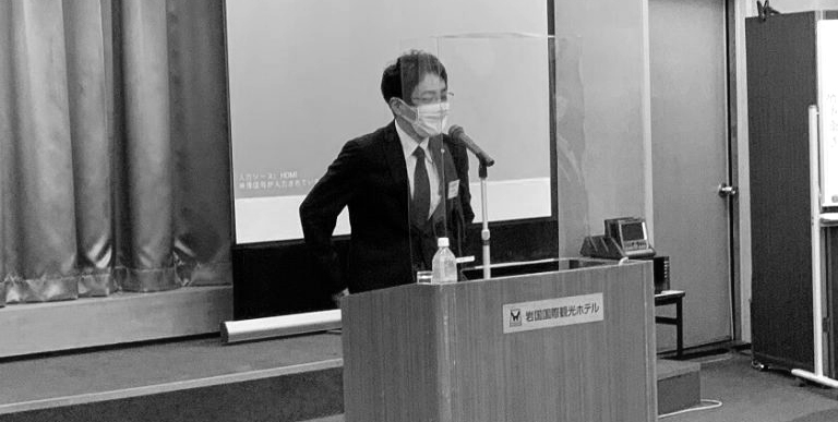 セミナー会場の演台で登壇し話をしている様子の写真(モノクロ、マスク着用)